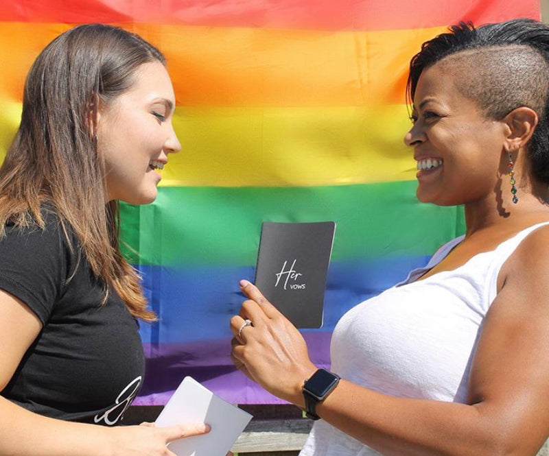 Two People Smiling at Each Other Holding Black and White Her Vow Books in Front of Rainbow Pride Flag