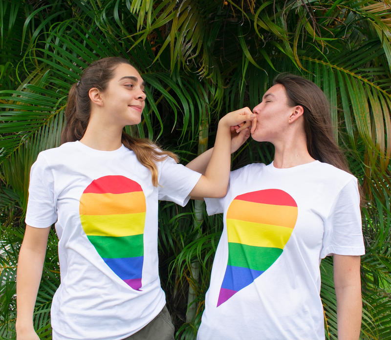 Two People Wearing Rainbow Pride Heart Tees Standing in Front of Palm Tree One Kissing the Other's Hand